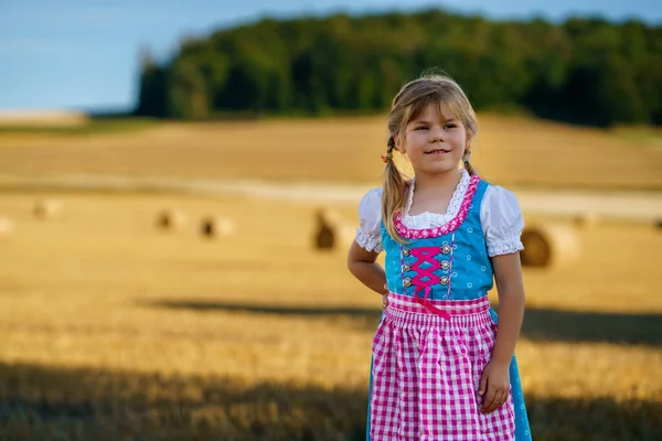 stock image Cute little kid girl in traditional Bavarian costume in wheat field. Happy child with hay bale during Oktoberfest in Munich. Preschool girl play at hay bales during summer harvest time in Germany