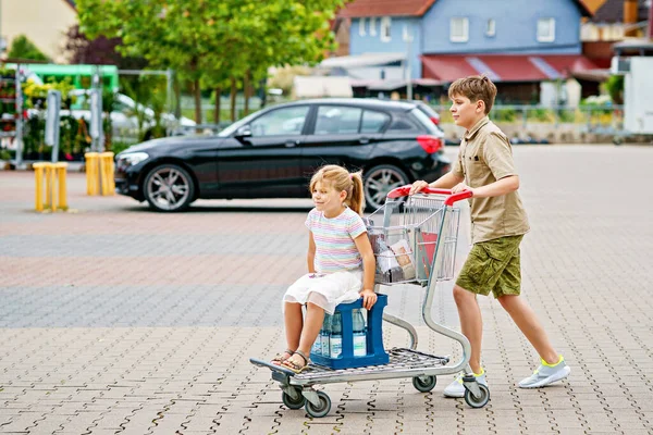 就学前の女の子が喜んで内部に座っている間 彼らは楽しい家族のショッピング旅行を楽しんでいるので 男の子はカートを押します スーパーマーケットの前にいる幸せな兄弟たち — ストック写真