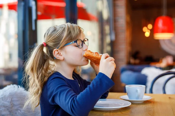 グラスを持つ愛らしい笑顔の女の子はカフェで朝食を持っています チョコレートを飲み パン屋さんのペストリークロワッサンやケーキを食べるメガネを持つ就学前の子供 幸せな子供たち 健康的な食べ物と食事 — ストック写真