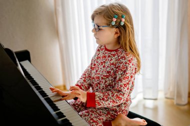 Little happy girl playing piano in living room. Cute preschool child with eye glasses having fun with learning to play music instrument