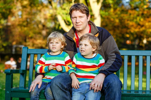 两个小孩子男孩和年轻的父亲坐在一起 穿着五颜六色的衣服在板凳上 可爱的健康的孩子 兄弟姐妹和他们的爸爸在秋季公园在温暖晴朗的一天乐趣 三幸福家庭 — 图库照片