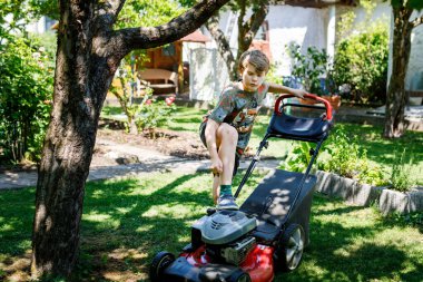 Çim biçme makineli mutlu çocuk. Bahçede çimleri biçen gülümseyen genç bir çocuğun portresi. Bahçe yazın çalışır. Çocuk yardım ediyor.