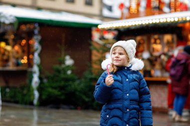 Noel pazarında şeker tezgahından şeker bastonuyla küçük şirin bir anaokulu kızı. Almanya 'nın geleneksel aile pazarında mutlu bir çocuk. Kar yağışı sırasında renkli kış kıyafetleri içinde anaokulu öğrencisi..
