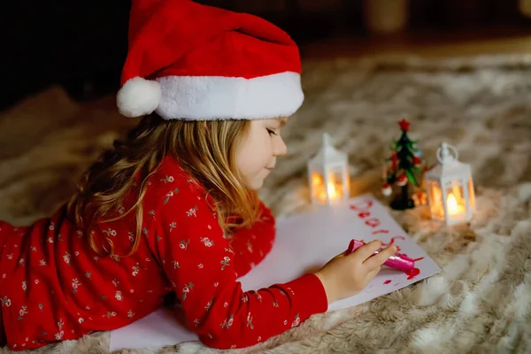 Little cute preschool girl in pajamas under Christmas tree, writing wish list letter to Santa Claus at home, indoors. Traditional Christian festival. Happy toddler child waiting for gifts on xmas