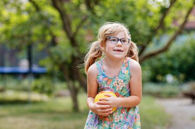Gözlüklü küçük anaokulu kızı dışarıda top oynuyor. Gülümseyen çocuk yakalayıp fırlatıyor, gülüyor ve spor yapıyor. Çocuklar ve çocuklarla aktif bir eğlence. Arka bahçede yaz günü.