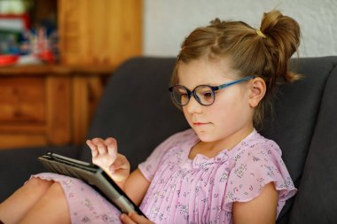 Tablet bilgisayarı elinde tutan pozitif küçük bir kız. Gözlüklü anaokulu çocuğu oyun oynuyor ve farklı uygulamalarla öğreniyor.