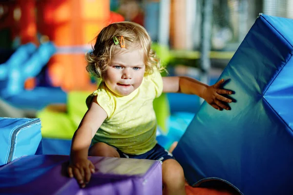 幸せな金髪幼児女の子と楽しんで保育園や保育園の屋内遊び場のスライドします 肯定的な面白い赤ちゃん子供が笑っています 健康的な女の子がスライドに登って — ストック写真