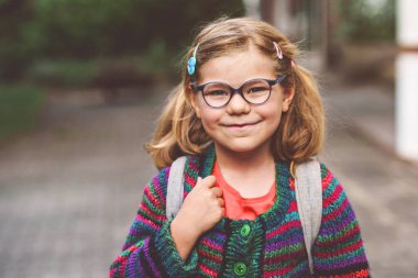 Okula giden sevimli, küçük bir anaokulu kızı. Gözlüklü, kreşe ve anaokuluna yürüyen sağlıklı, mutlu bir çocuk. Sırt çantalı gülen çocuk şehir caddesinde, açık havada. Okula geri dön.