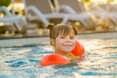 Gün batımında açık havuzda yüzen koruyucu yüzme elbiseli küçük bir anaokulu kızı. Çocuk havuzda yüzmeyi, su sıçratmayı, gülmeyi ve eğlenmeyi öğreniyor. Aile tatilleri