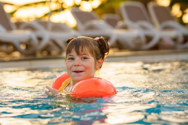 Gün batımında açık havuzda yüzen koruyucu yüzme elbiseli küçük bir anaokulu kızı. Çocuk havuzda yüzmeyi, su sıçratmayı, gülmeyi ve eğlenmeyi öğreniyor. Aile tatilleri