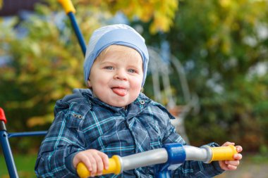 Dışarıda üç tekerlekli bisikletle oynayan şirin bir çocuk. Mutlu çocuk sonbahar parkında oynuyor. Küçük çocuk modaya uygun bir ceket giyiyor. Gülümseyen sarışın çocuk portresi. Sonbahar modası. Şık bir çocuk.