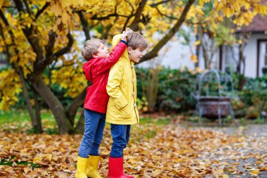 İki küçük arkadaş ve çocuklar sonbahar parkı rengarenk elbiseleriyle. Mutlu kardeşler, kırmızı ve sarı yağmurluklarla ve lastik botlarla eğleniyorlar. Aile dışarıda oynuyor. etkin boş zaman