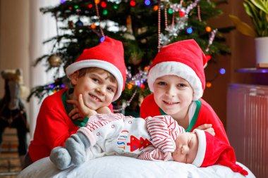 Bir hafta önce yeni doğan bebek kız ve iki kardeş Noel Baba şapkaları renkli garland ışıkları ile Noel ağacı yakınındaki çocuklar arka plan üzerinde evlat. Üç çocuk, yakın çekim mutlu aile Xmas kutluyor