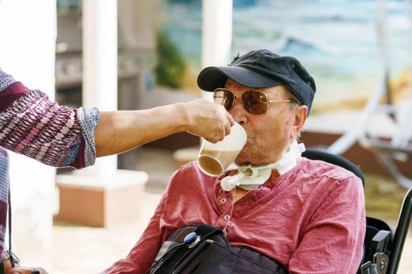 Senior man of 85 years in an electric wheelchair is sitting in a park. Nurse supporting man with drinking coffee, holding cup