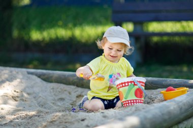 Dışarıda parkta kumlarda oynayan mutlu bir kız çocuğu. Güneşli sıcak yaz gününde bebek eğleniyor. Kum oyuncakları ve renkli kıyafetleri olan aktif bir çocuk.