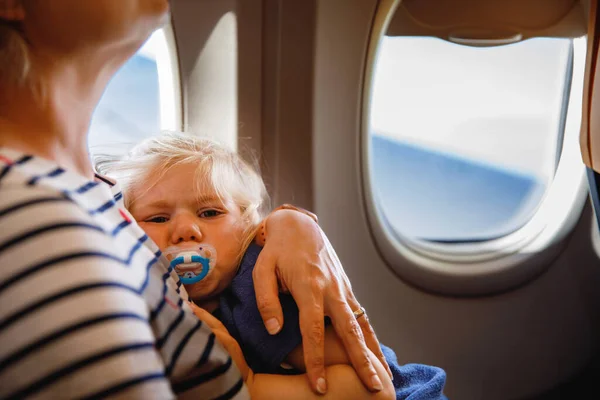 Woman traveling with little child by airplane. Sad tired toddler girl sitting with mum by aircraft window. Motherhood concept. Crying baby