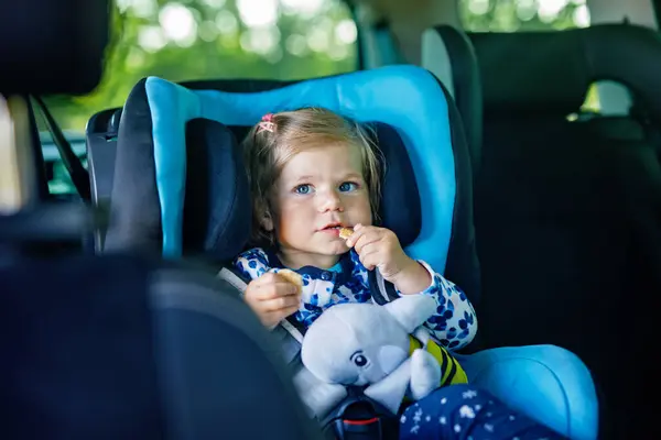 Adorable baby girl with blue eyes sitting in car safety seat. Toddler child going on family vacations and jorney. Smiling happy child during traffic jam, eating bisquit.