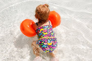 Gün batımında yüzme havuzunda oynayan koruyucu yüzme elbiseli küçük bir kız. Bebek havuzda yüzmeyi, su sıçratmayı, gülmeyi ve eğlenmeyi öğreniyor. Aile tatilleri