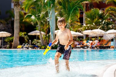 Küçük mutlu çocuk havuza atlıyor ve otel odasında aile tatillerinde eğleniyor. Suda su tabancasıyla oynayan sağlıklı bir çocuk. Gülen çocuk sıçrıyor.