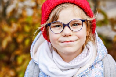 Açık havada gözlüklü ve kırmızı şapkalı sevimli bir anaokulu kızının portresi. Yeni mavi gözlük takan mutlu komik çocuk. Şehirde sonbahar günü