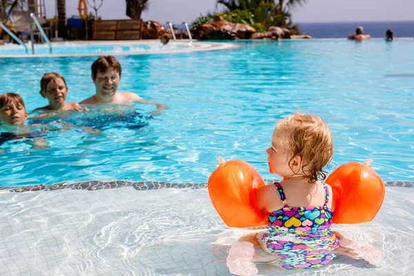 Cute happy little toddler girl, two school kid boys and father in the pool and having fun on family vacations in a hotel resort. Healthy children and man playing in water.