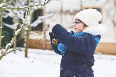 Little girl with glasses feeds birds on winter. Happy smiling preschool child hanging selfmade bird seed heart on tree. On cold snowy winter day