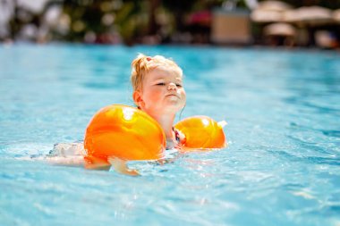 Gün batımında yüzme havuzunda oynayan koruyucu yüzme elbiseli küçük bir kız. Bebek havuzda yüzmeyi, su sıçratmayı, gülmeyi ve eğlenmeyi öğreniyor. Aile tatilleri