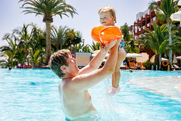 Cute happy little toddler girl and father in the pool and having fun on family vacations in a hotel resort. Healthy child and man playing in water. Baby daughter in colorful fashion swimsuit