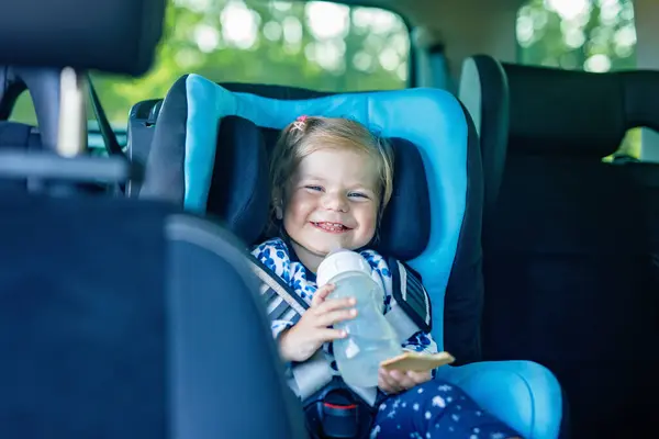Adorable baby girl with blue eyes sitting in car safety seat. Toddler child going on family vacations and jorney. Smiling happy child during traffic jam, drinking milk from bottle and eating bisquit.