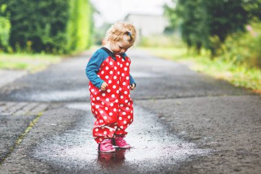 Yağmur botu ve pantolon giyen, yağmurda yürüyen, soğuk günlerde yağmur yağan küçük kız. Bebek su birikintisine atlamakla eğleniyor. Küçük çocuklarla açık hava eğlencesi ve etkinliği.