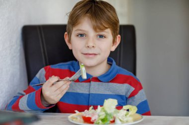 Mutlu çocuk domates, salatalık ve farklı sebzelerle taze salata yiyor. Sağlıklı bir çocuk evde ya da okul kantininde lezzetli ve taze yiyeceklerin tadını çıkarıyor.