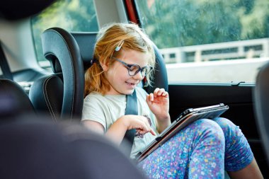 Küçük kız arabanın içinde tablet oynuyor. Aile tatile çıkıyor. Anaokulu çocuğu eğleniyor