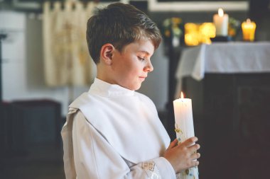 Küçük çocuk ilk kutsal komünyonunu alıyor. Vaftiz mumtutan mutlu çocuk. Katolik curch geleneği. Sunak yakınındaki bir kilisede beyaz geleneksel elbiseli çocuk