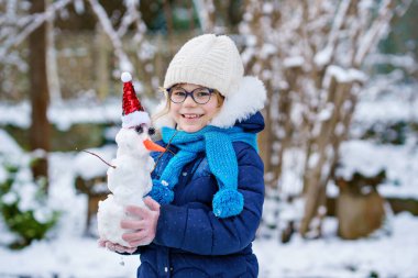 Cute little preschool girl with glasses making mini snowman. Adorable healthy happy child playing and having fun with snow, outdoors on cold day. Active leisure with children in winter.