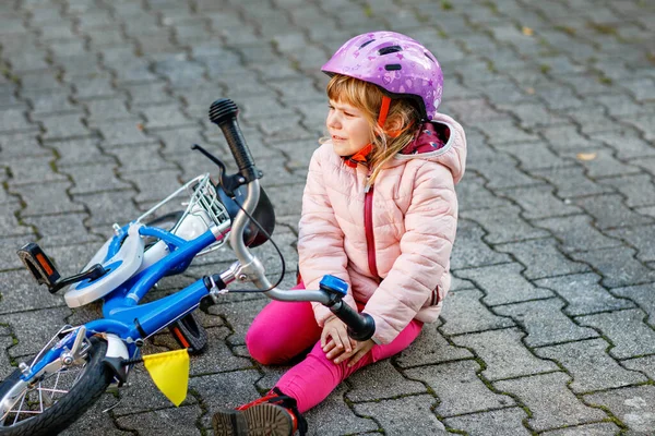 可爱的小女孩从自行车上摔下来 坐在地上 带着安全头盔的学龄前儿童在骑自行车时受伤 这让他很不高兴 有孩子的积极家庭休闲 — 图库照片