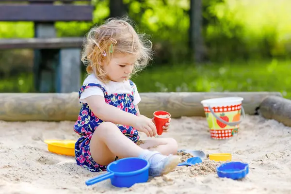 Dışarıda parkta kumlarda oynayan tatlı bir kız çocuğu. Kırmızı pantolonlu güzel bebek güneşli yaz gününde eğleniyor. Renkli kum oyuncakları olan çocuk. Sağlıklı aktif bebek dışarıda oyunlar oynuyor..