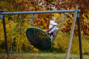Sonbaharda salıncakta sallanan mutlu kız çocuğu. Sonbaharda parkta oynayan, sallanan ve eğlenen küçük bir çocuk. Arka planda sonbahar ağaçları var. Okulun arka bahçesi