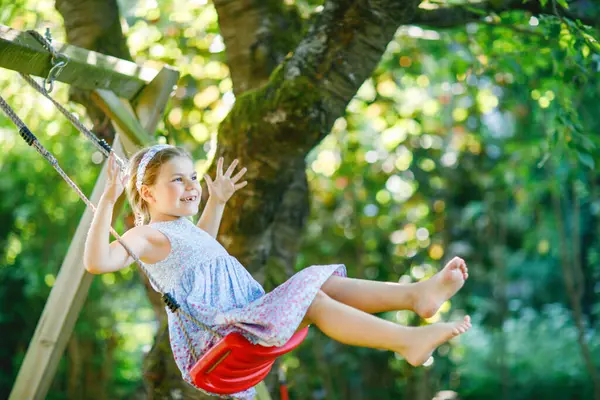 家庭菜園でスイングを楽しんで幸せな小さな就学前の女の子 晴れた春の日に咲く木々の下で揺れるかわいい健康的な子供 笑ったり泣いたり — ストック写真