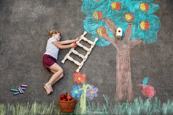 Cute little girl painting with colorful chalks apples harvest from apple tree on asphalt. Cute preschool child with having fun with chalk picture. Creative leisure for children, drawing and painting