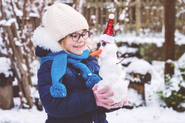 Cute little preschool girl with glasses making mini snowman. Adorable healthy happy child playing and having fun with snow, outdoors on cold day. Active leisure with children in winter.