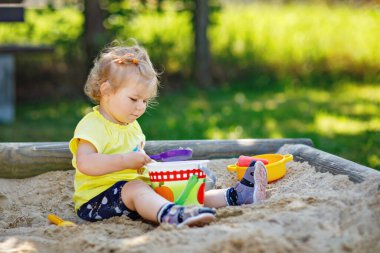 Happy toddler girl playing in sand on outdoor playground. Baby having fun on sunny warm summer sunny day. Active child with sand toys and in colorful fashion clothes