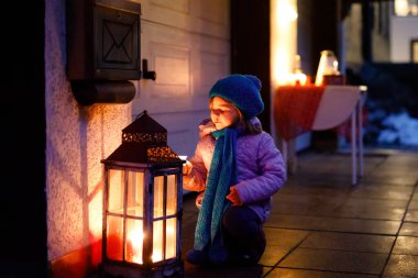 Elinde büyük bir mum feneri olan küçük tatlı bir kız. Evin arka bahçesindeki Noel süslemesinde mutlu çocuk. Aile tatili, aydınlanma, ışıklar ve mumlar.