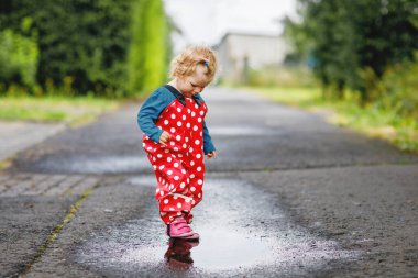 Yağmur botu ve pantolon giyen, yağmurda yürüyen, soğuk günlerde yağmur yağan küçük kız. Bebek su birikintisine atlamakla eğleniyor. Küçük çocuklarla açık hava eğlencesi ve etkinliği.