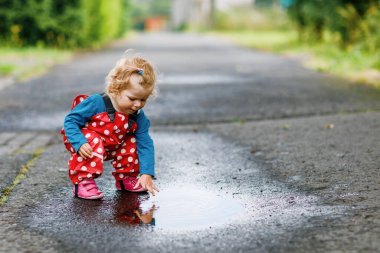 Yağmur botu ve pantolon giyen, yağmurda yürüyen, soğuk günlerde yağmur yağan küçük kız. Bebek su birikintisine atlamakla eğleniyor. Küçük çocuklarla açık hava eğlencesi ve etkinliği.