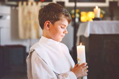 Küçük çocuk ilk kutsal komünyonunu alıyor. Vaftiz mumtutan mutlu çocuk. Katolik curch geleneği. Sunak yakınındaki bir kilisede beyaz geleneksel elbiseli çocuk