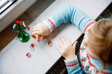 Küçük kız pencerenin kenarında oturuyor ve küçük cam Noel ağacını küçük Noel oyuncaklarıyla süslüyor. Mutlu sağlıklı çocuk geleneksel aile bayramını kutluyor. Sevimli bebek