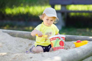 Dışarıda parkta kumlarda oynayan tatlı bir kız çocuğu. Güneşli sıcak yaz gününde güzel bir bebek eğleniyor. Kum oyuncakları ve renkli kıyafetlerle mutlu sağlıklı bir çocuk.