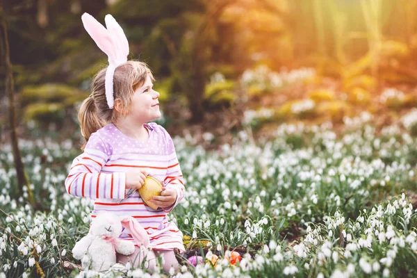晴れた日に春の森の中で卵狩りをするイースターバニーの耳を持つ少女 雪の花がたくさんあるかわいい幸せな子供 巨大なチョコレートの卵と色の卵 — ストック写真