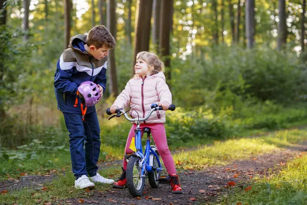 学校の少年 弟は小さな幼稚園の妹の少女の自転車のヘルメットを頭に置きました 幸せな子供のサイクリングを教え 自転車を学ぶことを楽しんでいます 屋外で活躍する兄弟姉妹 子供のアクティビティ — ストック写真