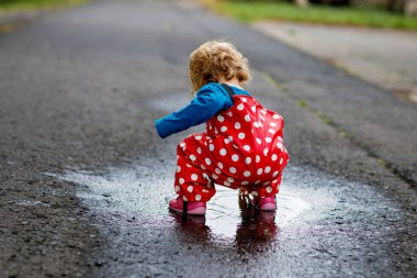 Yağmur botu ve pantolon giyen, yağmurda yürüyen, soğuk günlerde yağmur yağan küçük kız. Bebek su birikintisine atlamakla eğleniyor. Küçük çocuklarla açık hava eğlencesi ve etkinliği.